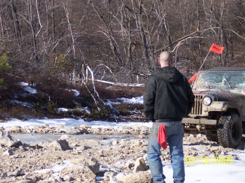 Man in jacket and jeans stands near a muddy Jeep Wrangler with an orange flag on a snowy trail