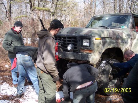 1995 Ford Bronco - Several individuals are gathered around the front of a camouflaged Ford Bronco, appearing to work on its tire in a wooded area.