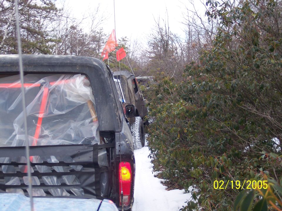 Back of a dark SUV with plastic covering the rear window, followed by another off-road vehicle with orange flags, driving on a snowy trail