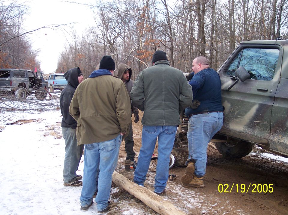 Group of men standing around a camouflage truck in a snowy, wooded area. Another SUV and truck are visible in the background
