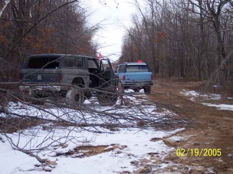 1995 Ford Bronco - Two modified SUVs are parked on a muddy and snowy trail surrounded by trees, with fallen branches obscuring the view.
