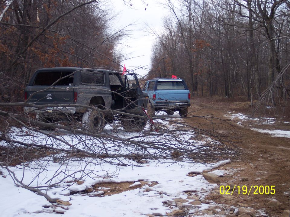 Two off-road SUVs, one dark and one blue, parked on a snowy, wooded trail with fallen branches in the foreground. The dark SUV has its door open