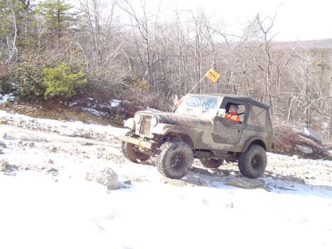 1995 Ford Bronco - Jeep Wrangler driving on a snowy, off-road trail with trees and rocks visible.
