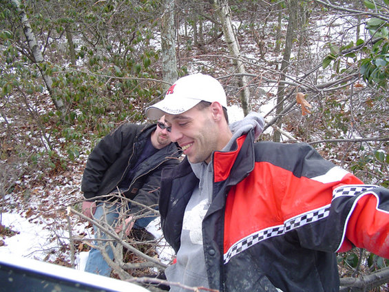 Two men in a snowy wooded area, one in a red and black jacket and white cap, the other in a dark jacket and sunglasses