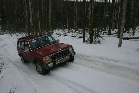 2003 Jeep TJ - A red Jeep Cherokee XJ navigates a snowy path through a forest, its body style indicating a 1984 model year.