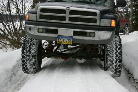 2003 Jeep TJ - A dark pickup truck with oversized tires navigates a snowy path, with another vehicle visible behind it.