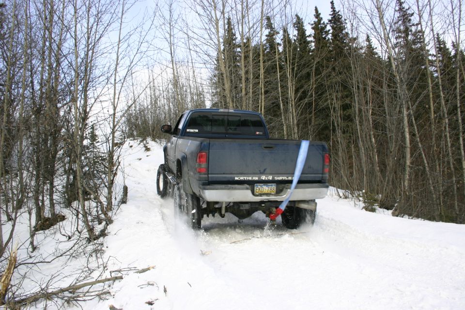 Dark blue pickup truck with a blue strap attached to the rear bumper driving on a snowy trail through trees