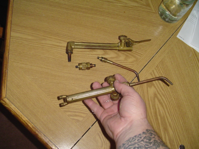 Hand holding a welding torch with a cutting attachment and a separate torch head on a wooden table