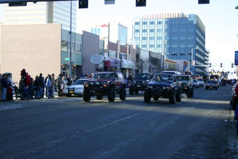 2003 Jeep TJ - Several modified off-road vehicles, including a 2003 Jeep TJ, participate in a parade on a city street.
