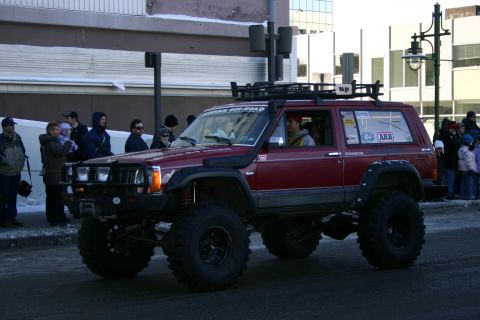 2003 Jeep TJ - A modified red Jeep Cherokee XJ is shown participating in a winter parade with onlookers on the sidewalk.
