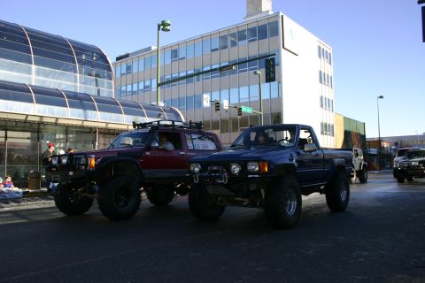 2003 Jeep TJ - Two modified off-road trucks participate in a parade on a sunny day in front of urban buildings.