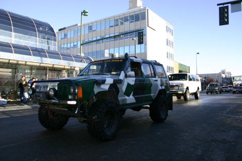 2003 Jeep TJ - A camouflaged Jeep Cherokee XJ with a snorkel and off-road tires drives down a street, with other vehicles visible in the background.