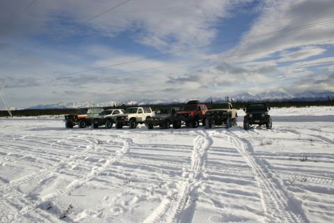 2003 Jeep TJ - A group of SUVs and trucks are lined up in a snow-covered field under a cloudy sky, with mountains visible in the distance.