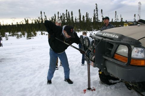2003 Jeep TJ - People work with a hi-lift jack on a vehicle in a snowy, wooded area, with power lines visible in the background.