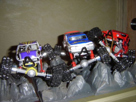 1992 Ford Bronco - A trio of remote-controlled monster trucks, each with a distinct color scheme, are positioned on a simulated rocky landscape.