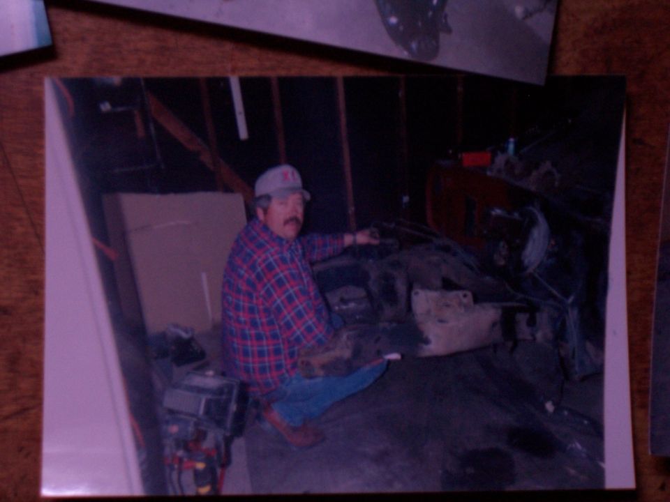 Old snapshot of man in plaid shirt kneeling next to a bare engine block in a wood-framed garage