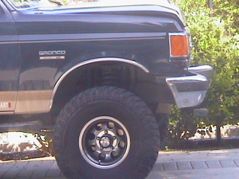 1991 Ford Bronco - Close-up view of the front quarter panel, wheel, and bumper of a dark blue 1991 Ford Bronco with tan trim and off-road tires.
