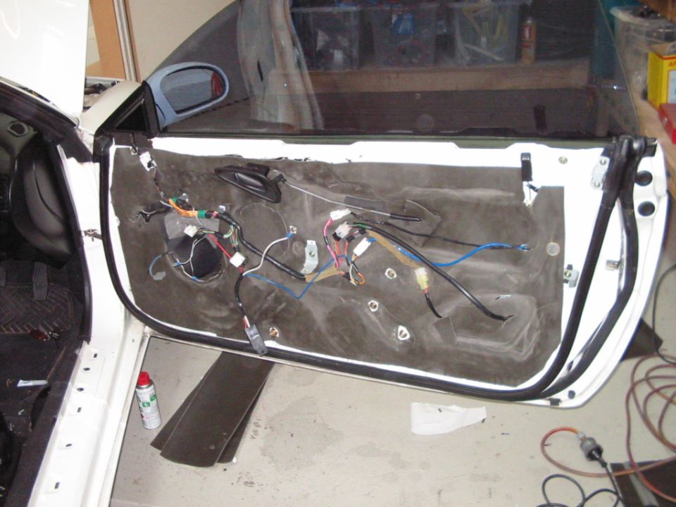 White car door interior with removed panel showing wiring, insulation, and speaker hole. A red spray can is on the floor