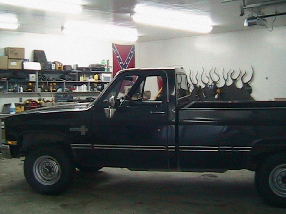 Black 1981 Chevrolet C/K Pickup truck, side view, in a garage with shelves and a Confederate flag on the wall