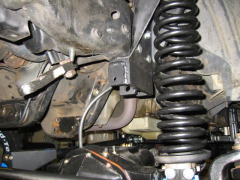 1992 Ford Bronco - Under-truck close-up of a welded steel track-bar bracket bolted to the frame beside a tall black front coil spring, with the weld beads and…