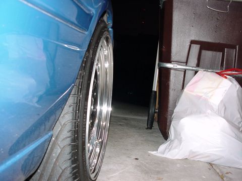 1998 Volkswagen Jetta - A close-up shot of a blue car's wheel and fender, with a storage bin and trash bag in the background.