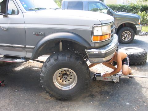 1992 Ford Bronco - A silver two-tone 1990s Ford Bronco with a suspension lift and large mud-terrain tires on chrome/aluminum wheels, front end raised on a Pro…
