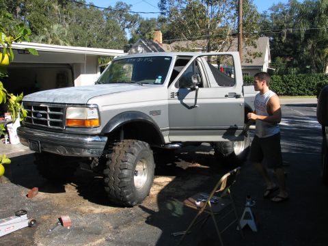 1992 Ford Bronco - A white two-tone Ford Bronco/F-series with a suspension lift and large mud-terrain tires on chrome wheels, parked on a driveway with the dr…
