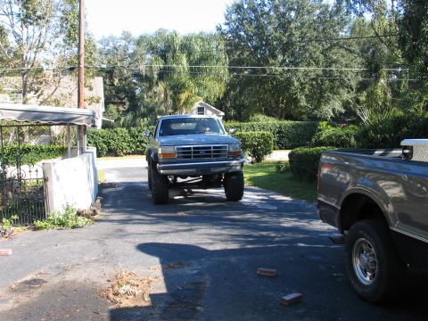1992 Ford Bronco - Head-on view of a light gray 1992-96 body-style Ford with a tall suspension lift and oversized tires rolling up a shaded driveway, with whi…