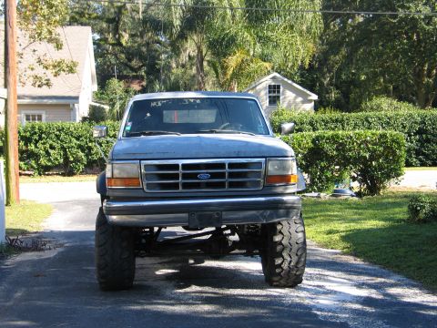 1992 Ford Bronco - Head-on front view of a lifted silver-gray Ford Bronco with a chrome grille and blue Ford oval, showing a solid front axle and tall mud tir…