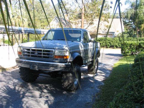 1992 Ford Bronco - A silver/gray lifted Ford pickup or SUV shown from the front three-quarter angle, sitting on large mud-terrain tires with chrome nerf/step…