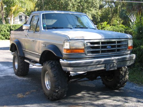 1992 Ford Bronco - A silver/gray Ford Bronco full-size SUV with a removable top (soft rear) shown from the 3/4 front angle, lifted on large off-road mud tires…