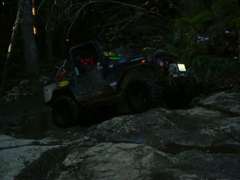 1990 Ford Bronco - A off-road vehicle navigates a challenging rocky path in a wooded area, with the Supermotors.net logo in the corner.