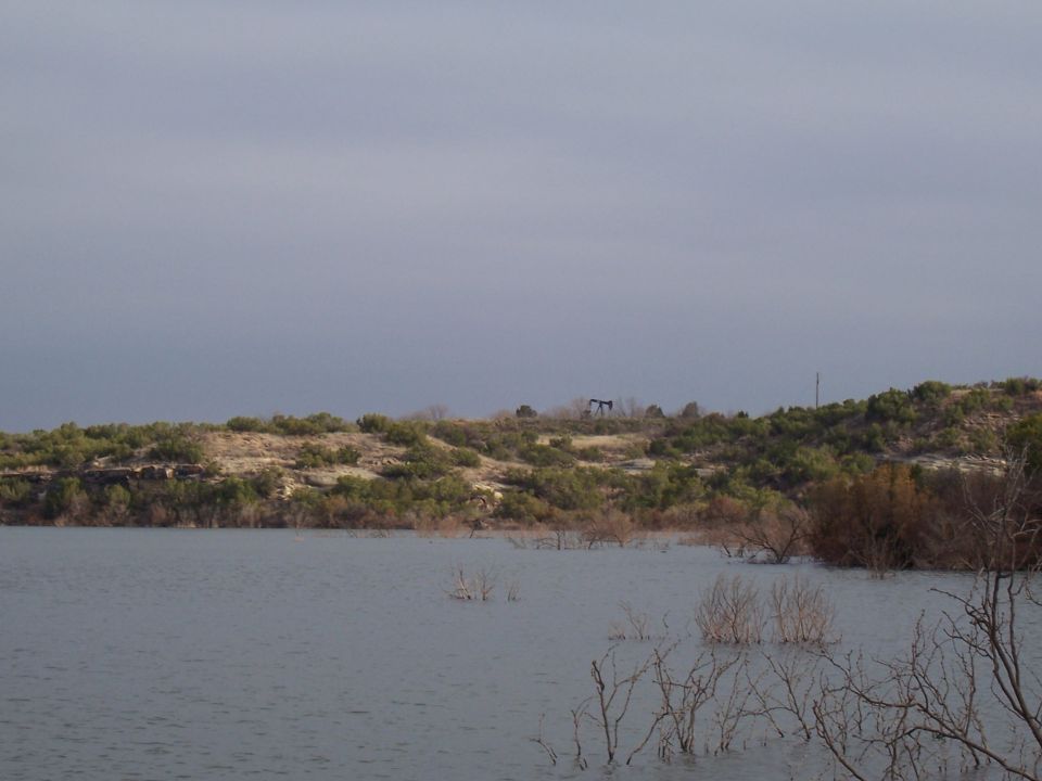 Reservoir with dead brush and an oil pumpjack on a scrub ridge under gray sky