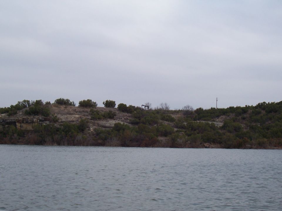 Lake view toward a brushy bluff with an oil pumpjack on the ridge under overcast sky