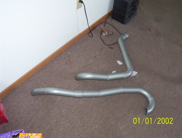 Two silver bent exhaust pipe sections lying on carpet near a baseboard, dated 01/01/2002