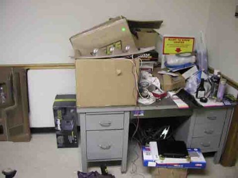 1988 Ford Bronco - A messy desk is piled high with boxes, papers, and various office supplies, with a yellow sign indicating a 'Drop Off Center'.