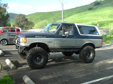 1989 Ford Bronco - A lifted, two-tone Ford Bronco with off-road tires is parked in a lot next to a red pickup truck.