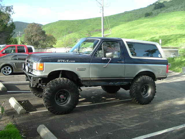 Lifted two-tone blue and silver Ford Bronco on big mud tires, side view in parking lot.