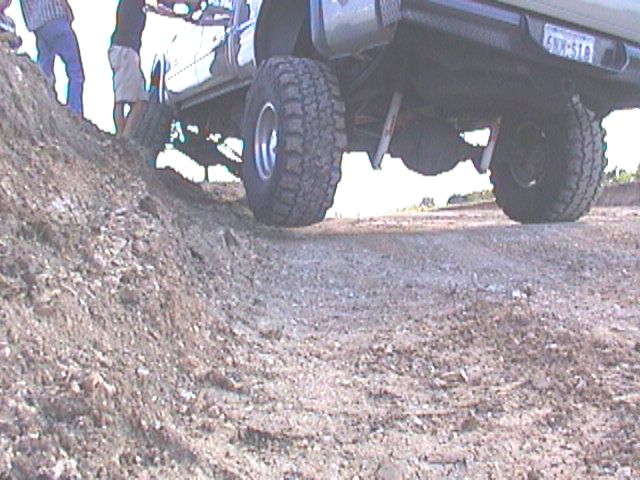 Off-road vehicle with large tires climbing a dirt mound, showing undercarriage and rear axle