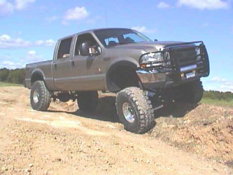 1989 Ford Bronco - A lifted pickup truck navigates a dirt track under a partly cloudy sky.