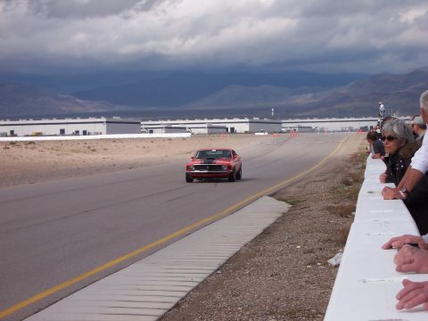 1971 Ford Mustang - A red muscle car speeds down a racetrack with spectators observing from the sidelines.