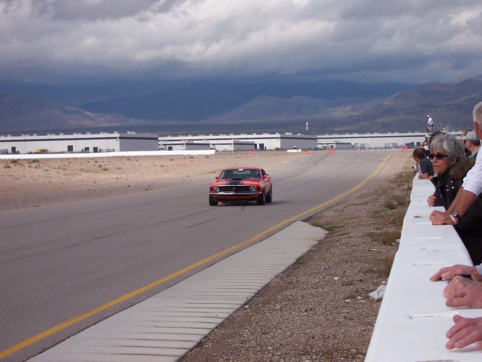 Red muscle car with black stripes driving on a track, spectators watch from the side