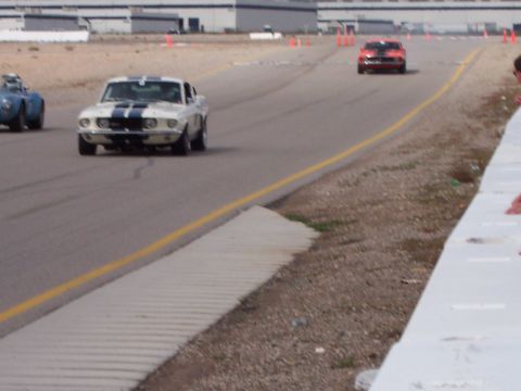 1971 Ford Mustang - A white 1969 muscle car with blue racing stripes navigates a turn on a race track, with other cars visible in the background.