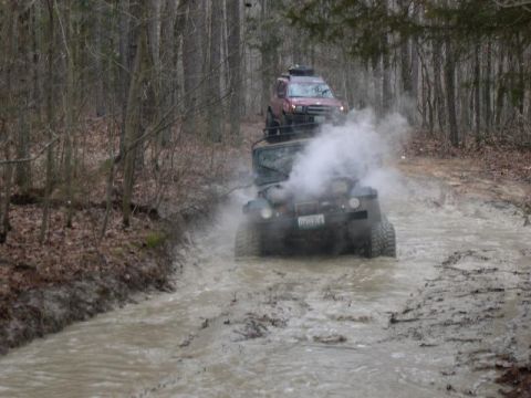 1999 Jeep TJ - Two off-road vehicles navigate a challenging muddy trail in a wooded area, one vehicle on top of the other.