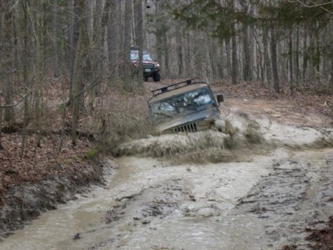 1999 Jeep TJ - A Jeep Wrangler TJ navigates a muddy trail, kicking up a large splash as another off-road vehicle follows behind.