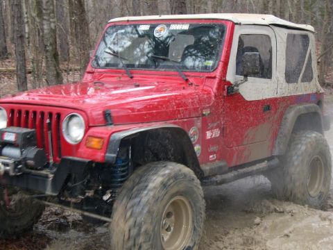 1999 Jeep TJ - This red Jeep Wrangler TJ navigates a muddy off-road course, its white soft top contrasting with the dirt.