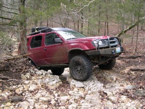 1999 Jeep TJ - This red SUV is navigating a rocky trail in a wooded environment, its off-road capabilities.