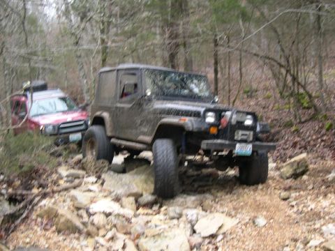 1999 Jeep TJ - A black 1999 Jeep TJ tackles a challenging rocky off-road path, with a red SUV following closely behind.