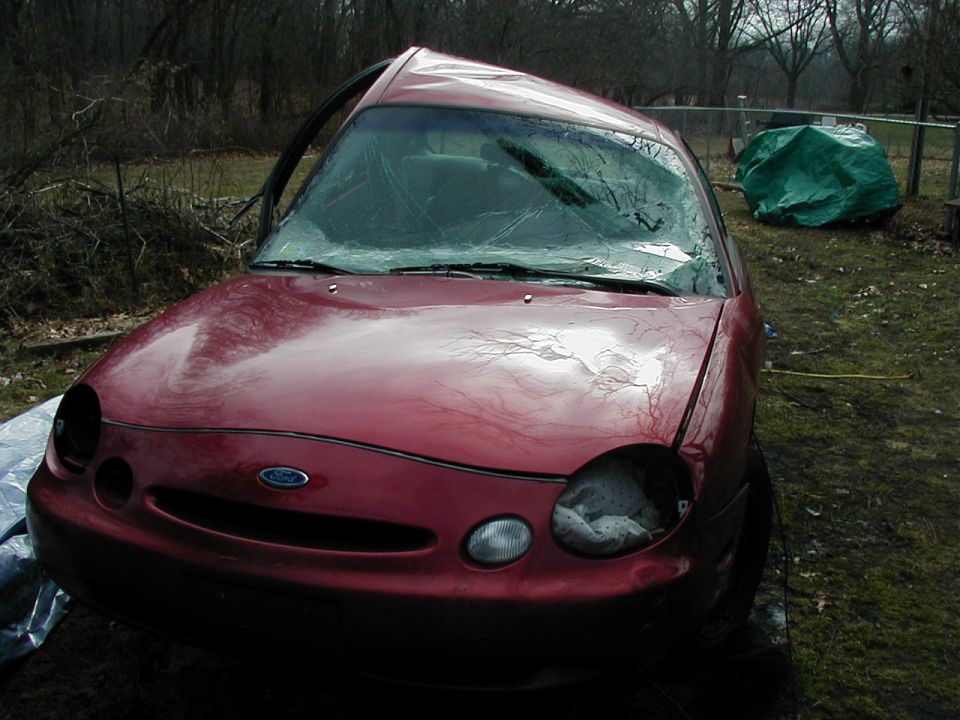 Red 1997 Ford Taurus with a crushed roof and broken headlights sits in a yard
