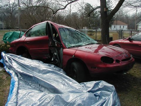 1997 Ford Taurus - A damaged red 1997 Ford Taurus sits in a yard with its driver's door open and windshield cracked, partially covered by a tarp.